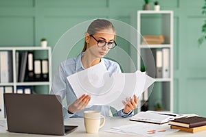 Concentrated businesswoman working with reports and documents, sitting with laptop at workplace in office interior