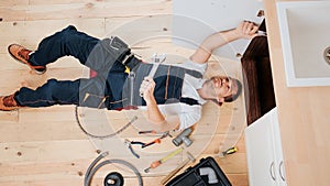 Concentrated busy plumber fixing on in kitchen under sink. He lying on floor. Guy hold wrench.