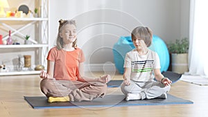 Concentrated Caucasian girl sitting on exercise mat with eyes closed as boy repeating yoga training after sibling. Wide