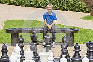 Concentrated child, thinking about his next move, sitting on a wooden bench during an outdoor chess game using life sized pieces