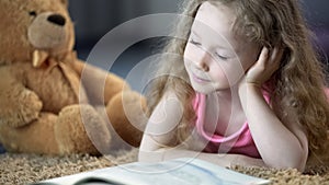 Concentrated cute girl reading book, lying on carpet, preschool education