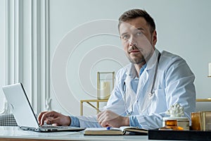 Concentrated doctor working with laptop at desk in office. Male doctor using laptop at desk in clinic