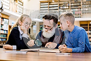 Concentrated elderly man teacher professor and his two little clever cute students reading book together. Grandfather