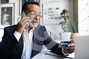 Concentrated european senior businessman in suit, glasses with laptop calls by phone, uses credit card