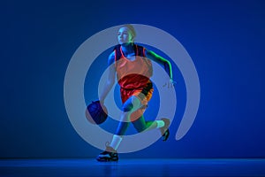 Concentrated female basketball athlete training, dribbling ball against blue studio background in neon light