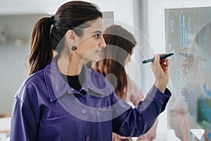 Concentrated, female, business worker strategizing with sticky notes on glass wall in modern office with colleague.