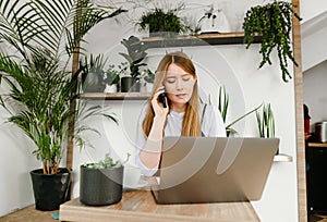 Concentrated freelance girl working in a cozy cafe with greenery and talking on the phone, looking into the screen. Business girl