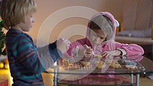 Concentrated girl decorating Christmas cookies with boy indoors. Portrait of cute Caucasian sister and brother preparing