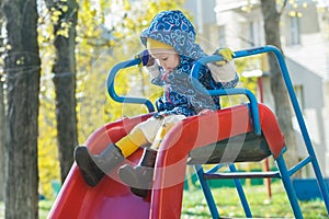 Concentrated girl in warm hooded jacket sliding down red plastic playground slide at yellow autumn tree leaves