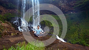 Concentrated guy meditates in yoga padma asana at waterfall