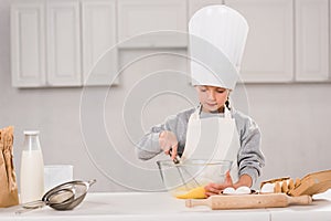 concentrated kid in chef hat whisking eggs in bowl at table