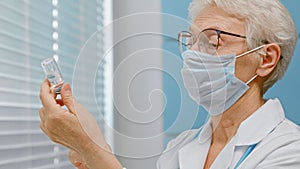 Concentrated lady doctor with protective mask fills syringe with vaccine from small vial near window in hospital