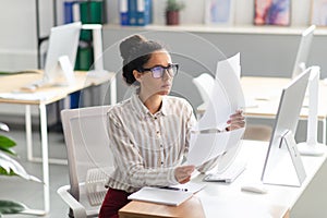Concentrated latin businesswoman working with documents and reports, sitting at workplace in office interior