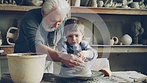 Concentrated little boy is learning to work with clay on throwing-wheel in pottery class in traditional workshop. His