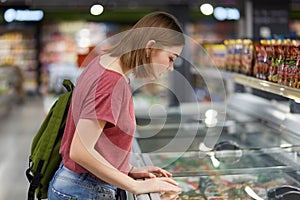 Concentrated lovely female teenager going to buy ice cream leans at fridge in supermarket, carries rucksack, dressed in casual clo