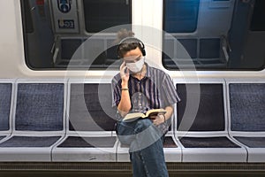 Concentrated masked millennial reading a book and listening to music in subway