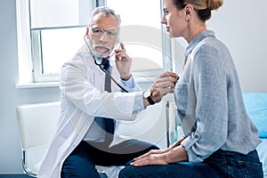 concentrated mature male doctor examining female patient by stethoscope