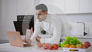 Concentrated Middle Eastern young man surfing Internet on laptop standing at countertop with ingredients for healthful