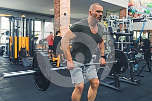 Concentrated millenial bald muscular guy lifting a heavy barbell using his triceps muscles. Indoor shot at a gym.