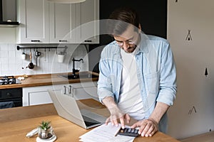 Concentrated millennial man planning monthly budget in kitchen.