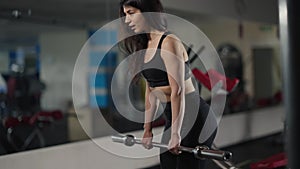 Concentrated motivated Middle Eastern woman lifting barbell bending forward indoors in gym. Side view portrait of