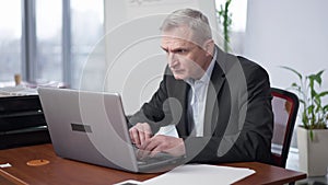 Concentrated senior Caucasian man sitting in office working online on laptop. Portrait of confident focused employee