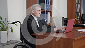 Concentrated senior disabled man in wheelchair typing on laptop keyboard in office. Portrait of confident Caucasian