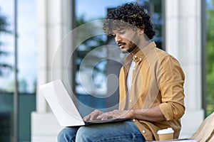 Concentrated and serious young Indian man using a laptop while sitting on a bench outside