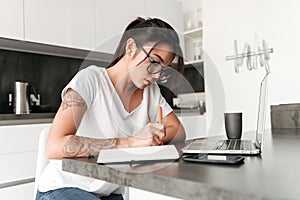 Concentrated serious young woman using laptop computer writing notes.