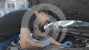 Concentrated service man examining car engine in open hood. Side view of young Caucasian auto mechanic fixing or tuning