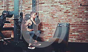Concentrated sporty girl exercising squatting with barbell at  smith machine against brick wall in gym.