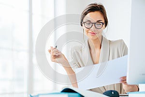 Concentrated successful businesswoman looks attentively in paper, studies terms of contract, holds pen, writes in documentations,