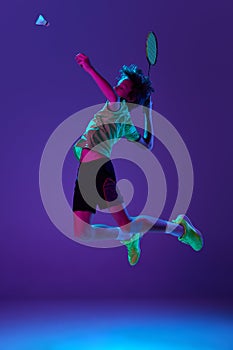 Concentrated teen boy in uniform playing badminton, hitting shuttlecock in a jump over blue purple background in neon