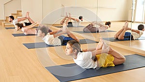 Concentrated teen girl doing back bending asana Dhanurasana known as Bow Pose during yoga workout at gym with family