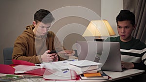 Concentrated teenage Asian boy showing laptop screen to his Caucasian groupmate using smartphone. Two handsome male