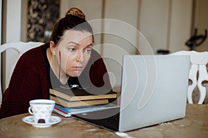Concentrated tired woman reading info, drink coffee, using laptop, remote job at home. Work problems at freelance office