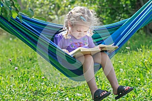 Concentrated two years old girl reading opened book on hanging hammock in green summer garden outdoors