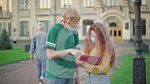 Concentrated university students in face masks examining homework outdoors as blurred man and woman greeting each other