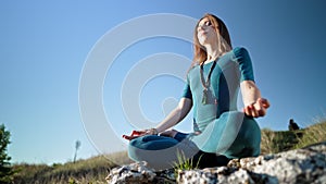 Concentrated woman in lotus pose doing meditation on high cliff above water. Calm yoga concept, religion, zen, peaceful