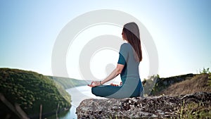 Concentrated woman in lotus pose doing meditation on high cliff above water. Calm yoga concept, religion, zen, peaceful