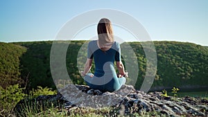 Concentrated woman in lotus pose doing meditation on high cliff above water. Calm yoga concept, religion, zen, peaceful