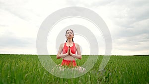 Concentrated woman in lotus pose doing meditation with namaste in green field. Calm yoga concept, religion, zen