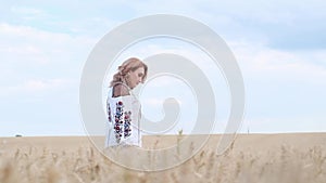 A concentrated woman in Ukrainian clothes walks through a wheat field. Blue sky background.