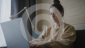 Concentrated woman works on laptop. A woman sits in the living room on a sofa near the window. Female freelancer uses modern PC.