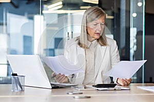 Concentrated and worried scary gray-haired businesswoman sitting in the office at the desk, reviewing and holding