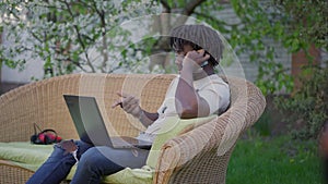 Concentrated young African American man talking on the phone sitting in spring summer garden with laptop. Portrait of