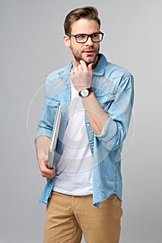 Concentrated young bearded man wearing glasses dressed in jeans shirt holding laptop isolated over grey studio