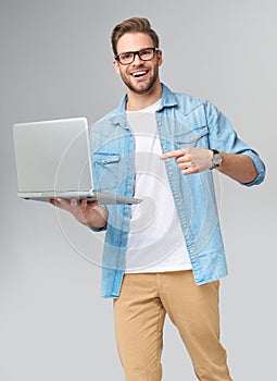 Concentrated young bearded man wearing glasses dressed in jeans shirt holding laptop isolated over grey studio