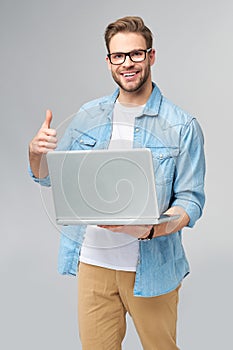 Concentrated young bearded man wearing glasses dressed in jeans shirt holding laptop isolated over grey studio