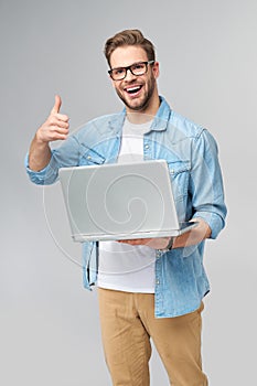 Concentrated young bearded man wearing glasses dressed in jeans shirt holding laptop isolated over grey studio